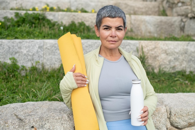 Free photo portrait of woman with grey hair after training in park. woman in sportive clothes holding wrapped mat and thermos. sport, nature concept