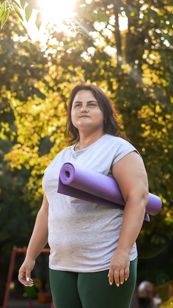 Free photo portrait of a woman with overweight in a tracksuit on a sports field in a park holding a yoga mat