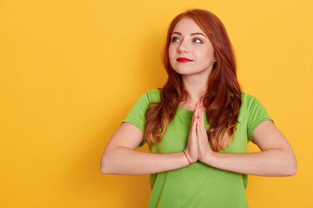 Portrait of woman with red hair in basic green t-shirt keeping palms together and praying, looking aside isolated, beautiful lady