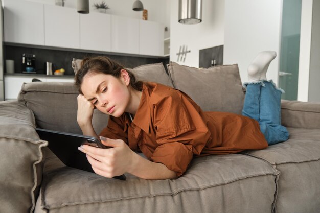 Free photo portrait of woman with sad face lying on couch at home resting in living room with digital tablet