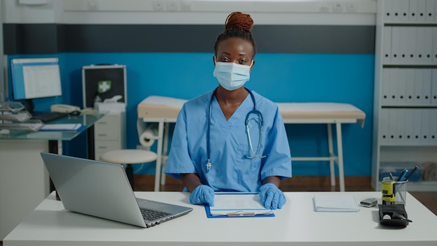 의료 클리닉에 앉아 있는 동안 얼굴 마스크와 제복을 입은 의료 보조원으로 일하는 여성의 초상화. 병원 방에서 카메라를 보고 있는 노트북과 파일이 있는 책상에 있는 간호사