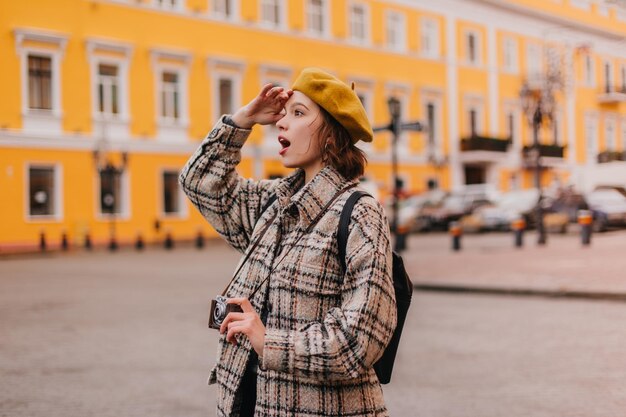 Portrait of woman in yellow beret with retro camera in her hands Girl in plaid coat looks at magnificent European buildings with surprise and delight