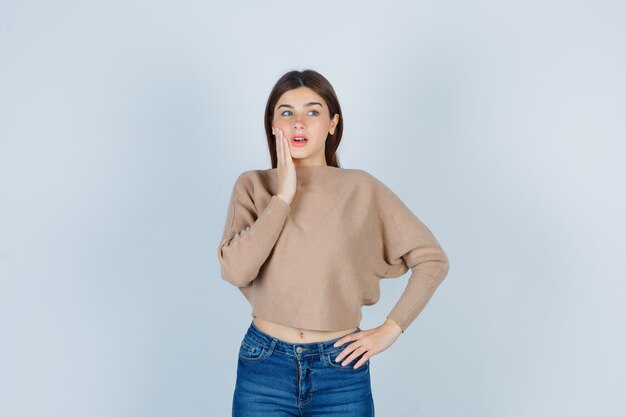 Portrait of wonderful lady keeping finger on cheek, opening mouth in sweater, jeans and looking astonished front view