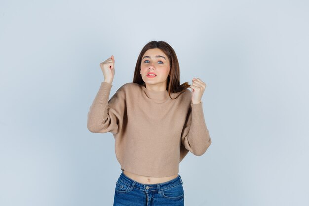 Portrait of wonderful lady raising clenched fists in sweater, jeans and looking furious front view