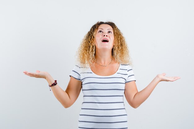 Portrait of wonderful lady spreading palms aside while looking up in striped t-shirt and looking surprised front view