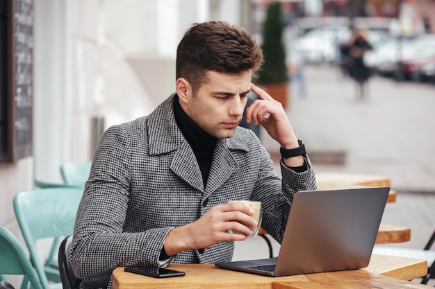 Portrait of working man sitting with silver laptop in cafe outside, drinking americano from glass