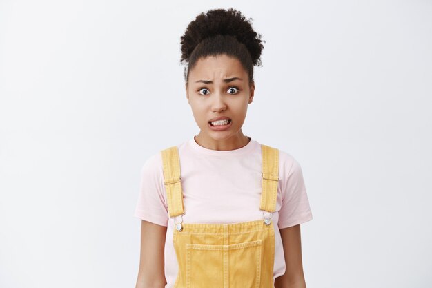 Portrait of worried insecure attractive woman with dark skin and curly hair in bun hairstyle, clenching teeth, making guilty expression