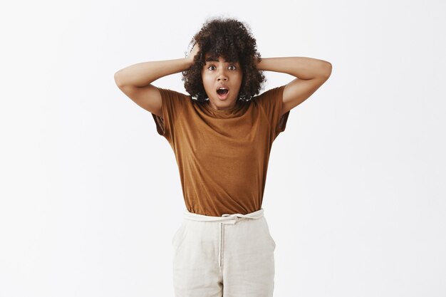 Portrait of worried insecure and stunned girl with afro hairstyle holding hands on head gasping and opening mouth being in hopeless expression