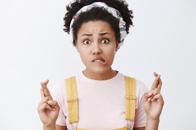 Portrait of worried intense charming dark-skinned female in dungarees and headband, biting lip and crossing fingers in hope gesture