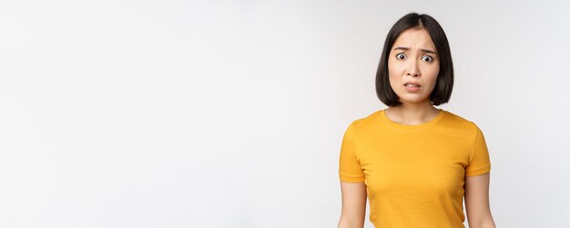 Portrait of worried korean girl looking concerned at camera standing in yellow tshirt over white bac