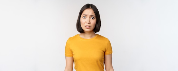 Portrait of worried korean girl looking concerned at camera standing in yellow tshirt over white background