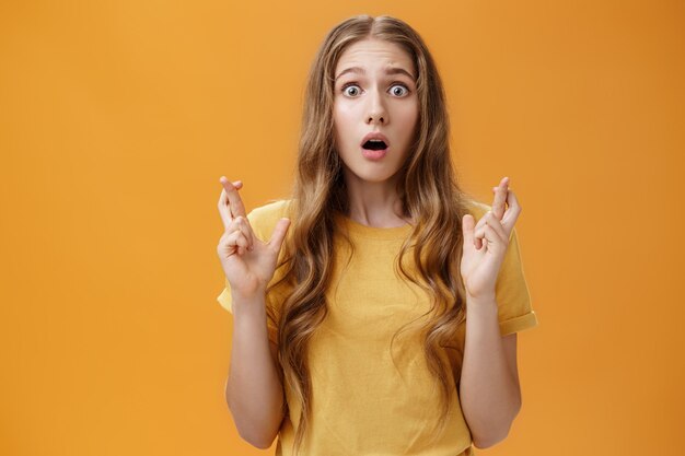 Portrait of worried silly young cute woman with wavy natural hairstyle crossing fingers for good luck opening mouth looking with hope and faith at camera praying, making wish over orange wall.