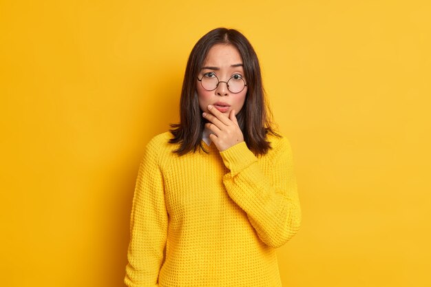 Portrait of worried surprised Asian woman holds chin looks concerned wears transparent glasses and sweater.