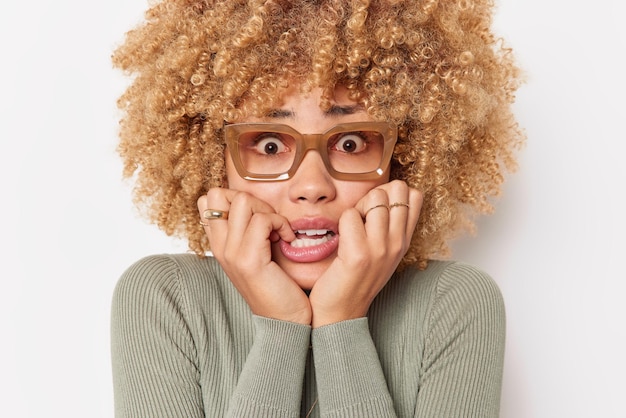 Foto gratuita ritratto di donna preoccupata con capelli ricci e folti morde le unghie delle dita sente la pressione e lo stress ha paura di cosa spaventosa indossa occhiali maglione casual isolato su sfondo bianco. ansia delle persone