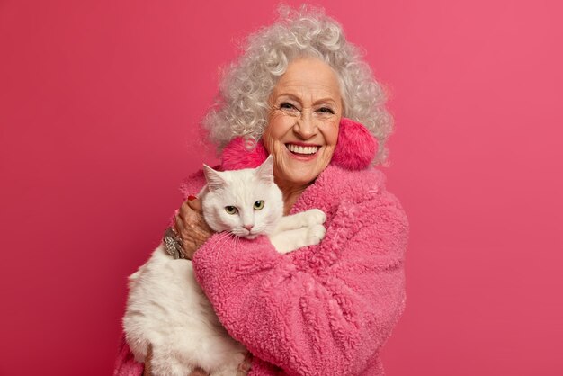 Portrait of wrinkled grandmother holds white cat on hands, stays at home during pandemic outbreak, wears fluffy earrings, soft robe, going to feed pet, isolated on pink wall. Woman on pension