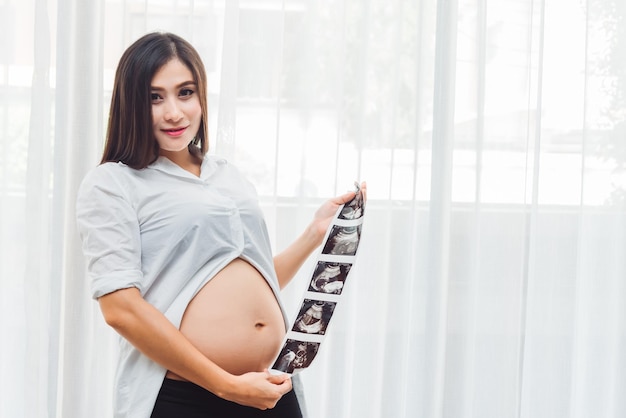 Free photo portrait of young adult asian pregnant woman holding ultrasound scan photo with happy