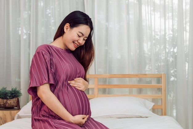 Portrait of young adult asian pregnant woman touching her belly with love at home