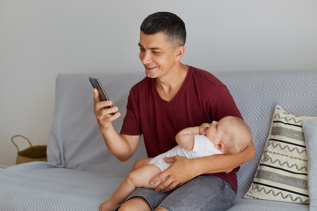 Portrait of young adult Caucasian man wearing maroon casual style t shirt with cute child in white attire, father holding his baby while using his phone while sitting on sofa in light room.