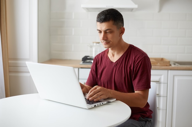 Portrait of young adult man wearing casual style maroon t shirt, freelancer working online from home, sitting at the table in kitchen, looking concentrated at notebook display.