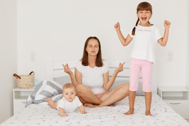 그녀의 어린 딸들이 엄마 근처에서 노는 동안 요가 자세로 아침 운동을 하는 다른 젊은 성인의 초상화, 주먹을 꽉 쥐고 서 있는 미취학 아동, 배에 누워 있는 유아.