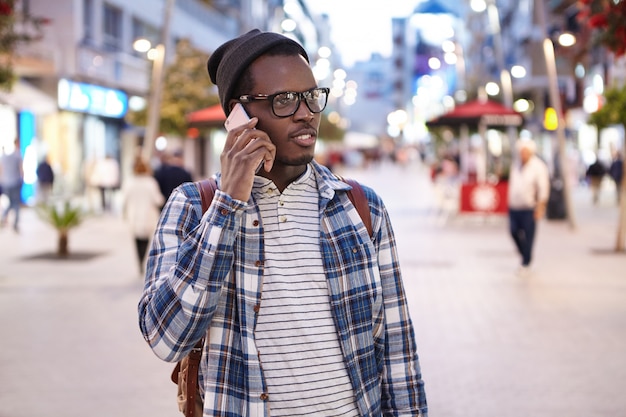 Portrait of young African American male wearing stylish clothing and accessories talking on smartphone on his way home