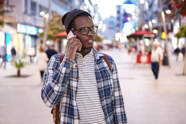 Portrait of young African American male wearing stylish clothing and accessories talking on smartphone on his way home