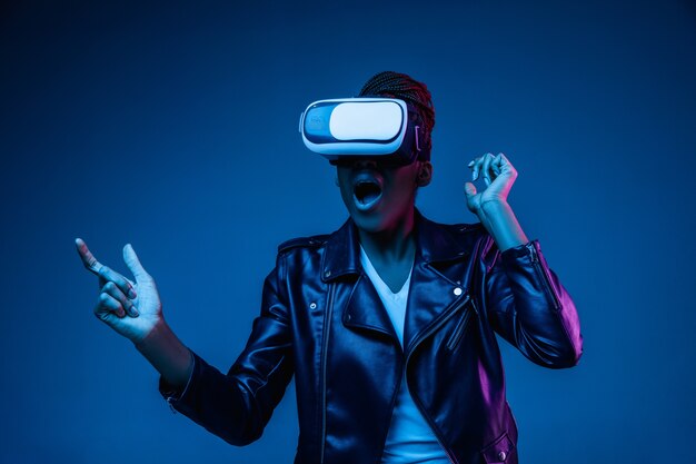 Portrait of young African-american woman playing in VR-glasses in neon light on blue.