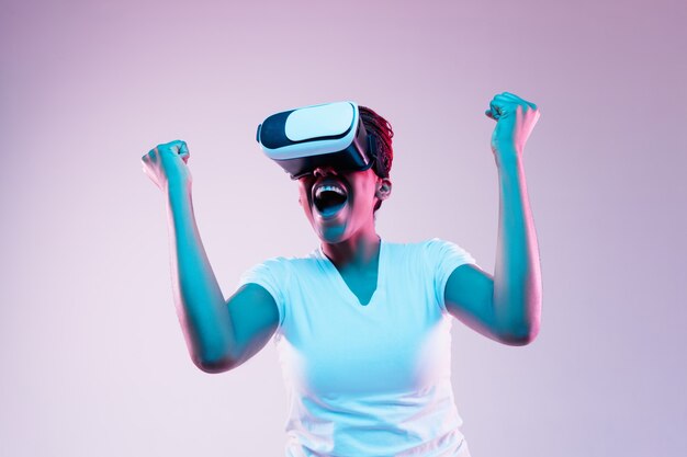 Portrait of young african-american woman's playing in VR-glasses in neon light on gradient background. Concept of human emotions, facial expression, modern gadgets and technologies. Look like winner.