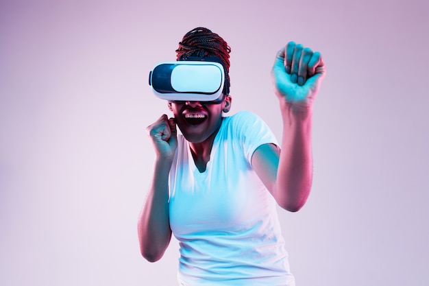 Portrait of young african-american woman's playing in VR-glasses in neon light on gradient background