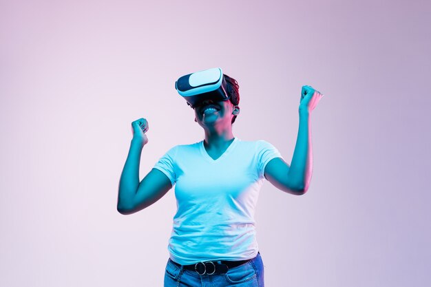 Portrait of young african-american woman's playing in VR-glasses in neon light on gradient.