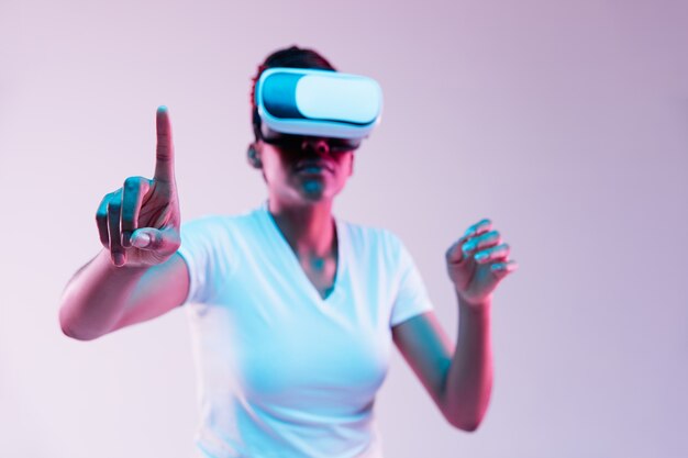 Портрет молодой афро-американской женщины, играющей в VR-очках в неоновом свете на градиенте.
