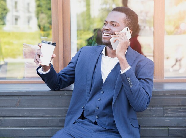 Портрет молодого африканского бизнесмена держа кофейную чашку на вынос говоря на мобильном телефоне