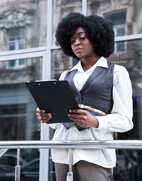 Free photo portrait of a young african businesswoman standing in front of railing holding clipboard