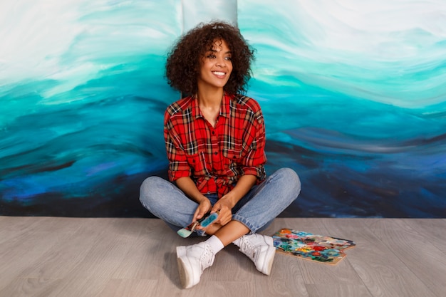 Portrait of  young african student sitting with amazing abstract acrylic hand drawn artwork at the studio.