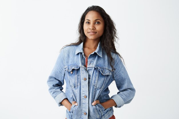 Portrait of young Afro-American girl with dark skin wearing denim jacket and red t-shirt  with serious expression, standing with her hands in pockets. Dark-skinned female model with d