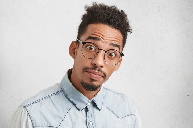 Portrait of young Afro American man with bugged eyes, has trendy hairdo, mustache and beard, looks puzzled