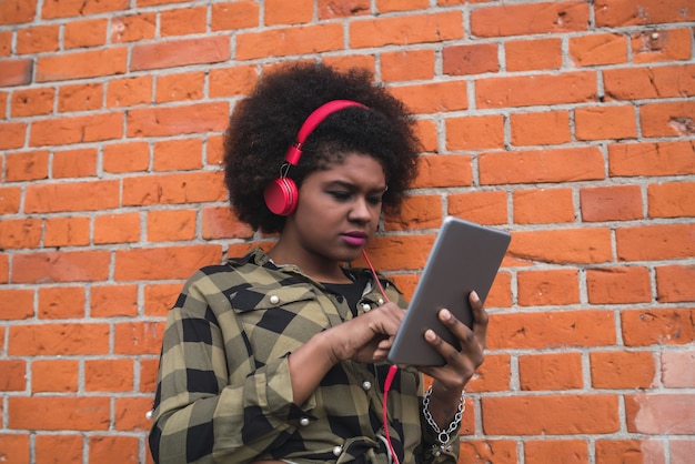 Портрет молодой афро-американской женщины, использующей ее цифровой планшет с красными наушниками на открытом воздухе. Концепция технологии.