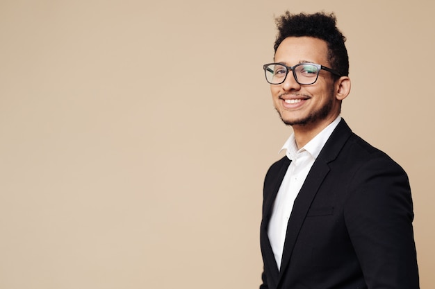 Portrait of young afro businessman wearing shirt, black suit, glasses and looking at front while standing at beige wall