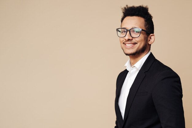 Portrait of young Afro businessman wearing shirt, black suit, glasses and looking at front while standing at beige wall