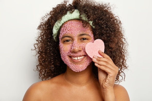 Portrait of young Afro female model holds heart shaped sponge near face covered with salt granules, smiles broadly, has white teeth with little gap, stands naked, expresses positive emotions