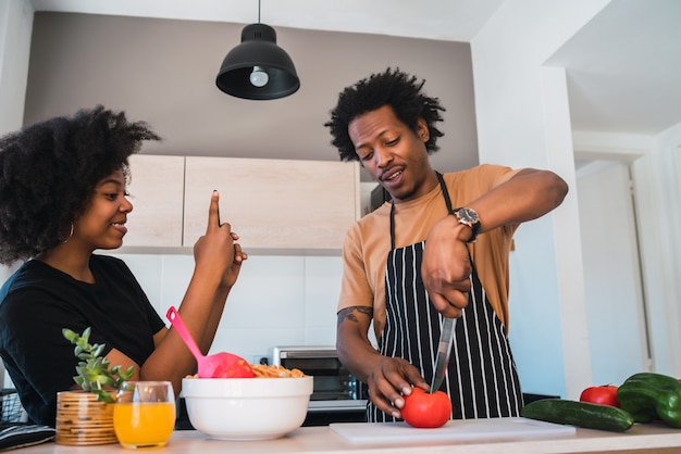 Portrait of young afro woman taking photo of her husband while he prepares dinner. Relationship, cook and lifestyle concept.