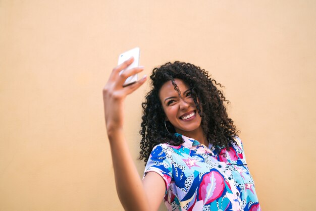 노란색 벽에 그녀의 mophile 전화 selfies를 복용하는 젊은 아프리카 여자의 초상화. 기술 개념.
