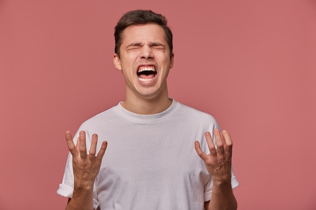 Free photo portrait of young angry guy in blank t-shirt, hears bad news and looks evil, stands on pink and screaming with unhappy expression.