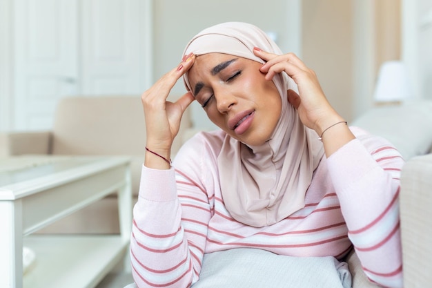 Portrait of a young Arabic Muslim woman sitting on the couch at home with a headache and pain Woman with hijab suffering from chronic daily headaches Sad woman holding her head because sinus pain