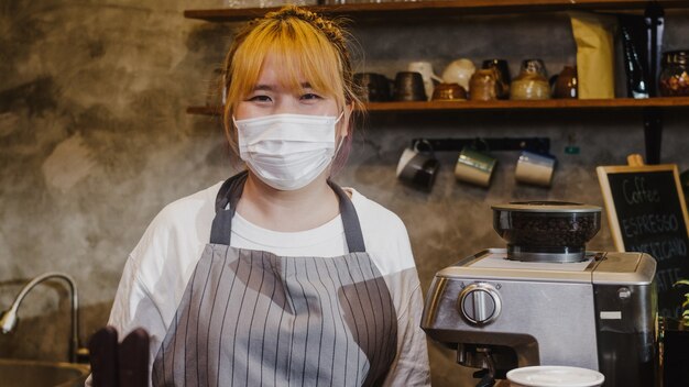 Портрет молодой официантки девушки Азии носить медицинскую маску для лица, чувствуя счастливую улыбку, ожидая клиентов после блокировки в городском кафе.