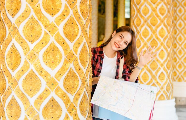 Portrait Young Asian backpacker female standing and hold paper map in hand at beautiful Thai temple, she smile during waving hand 