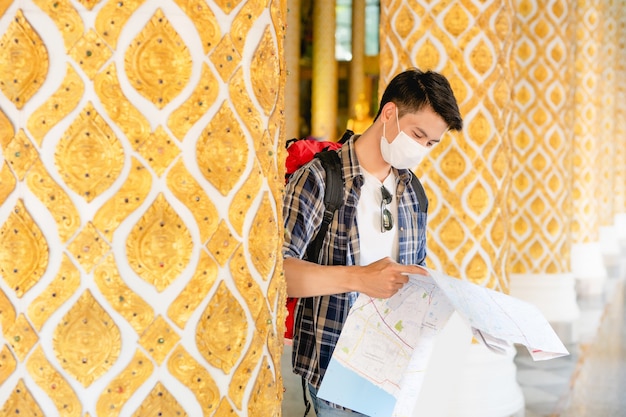 Portrait Young Asian backpacker male in face mask standing and checking direction on paper map in hand at beautiful Thai temple