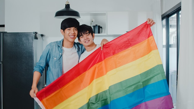 세로 젊은 아시아 게이 커플 집에서 무지개 깃발을 보여주는 행복 느낌. 아시아 LGBTQ + 남성은 아침에 집에서 현대 부엌에서 포옹하면서 카메라를 찾고 이빨 미소 휴식.