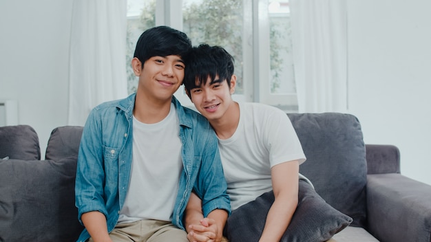 세로 젊은 아시아 게이 커플 집에서 웃 고 행복 한 느낌. 아시아 LGBTQ 남성은 아침에 집에서 거실 소파에 누워 카메라를 찾고 이빨 미소 휴식.