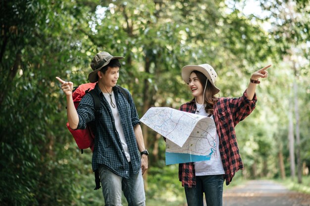 Portrait Young asian handsome man with backpack and trekking hat and pretty girlfriend standing and checking direction on paper map while walking on forest trail, backpack travel concept